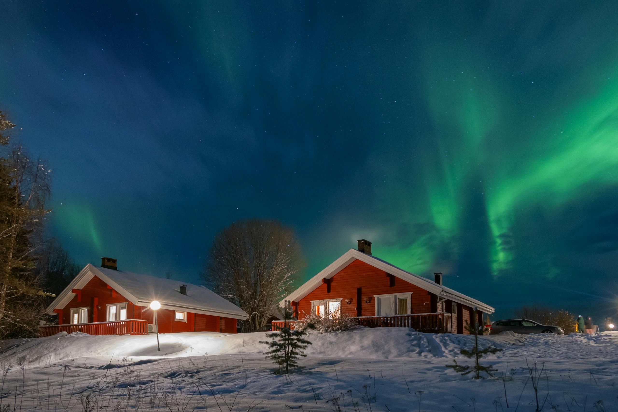 Red wooden cabins under the vibrant aurora borealis in a snowy Finnish landscape.